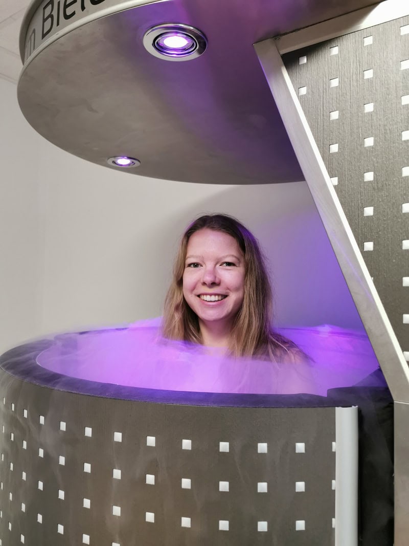 Frau lächelt in einer kryotherapeutischen Kammer, umgeben von lila Licht und Nebel.