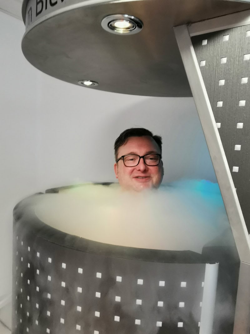 Ein lächelnder Mann in einer Kryokammer, umgeben von Nebel und farbigem Licht.