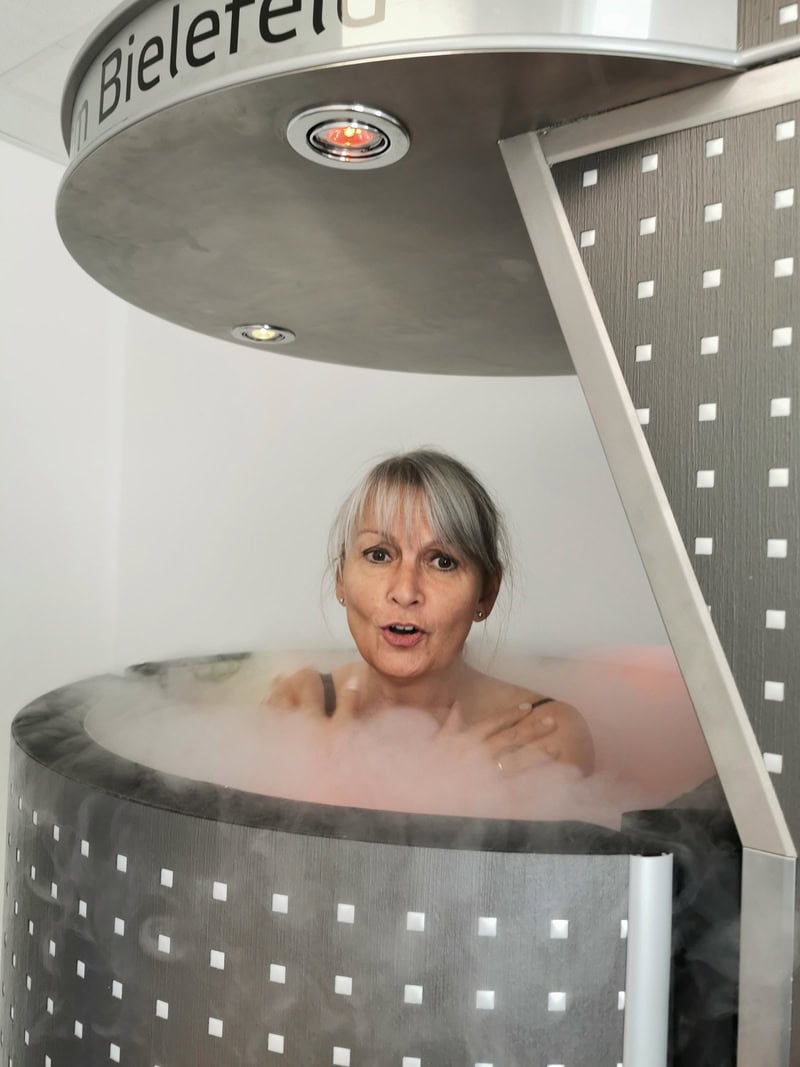 Ältere Frau mit grauem Haar in Kryosauna, überrascht, umgeben von Nebel in metallischer Kammer.