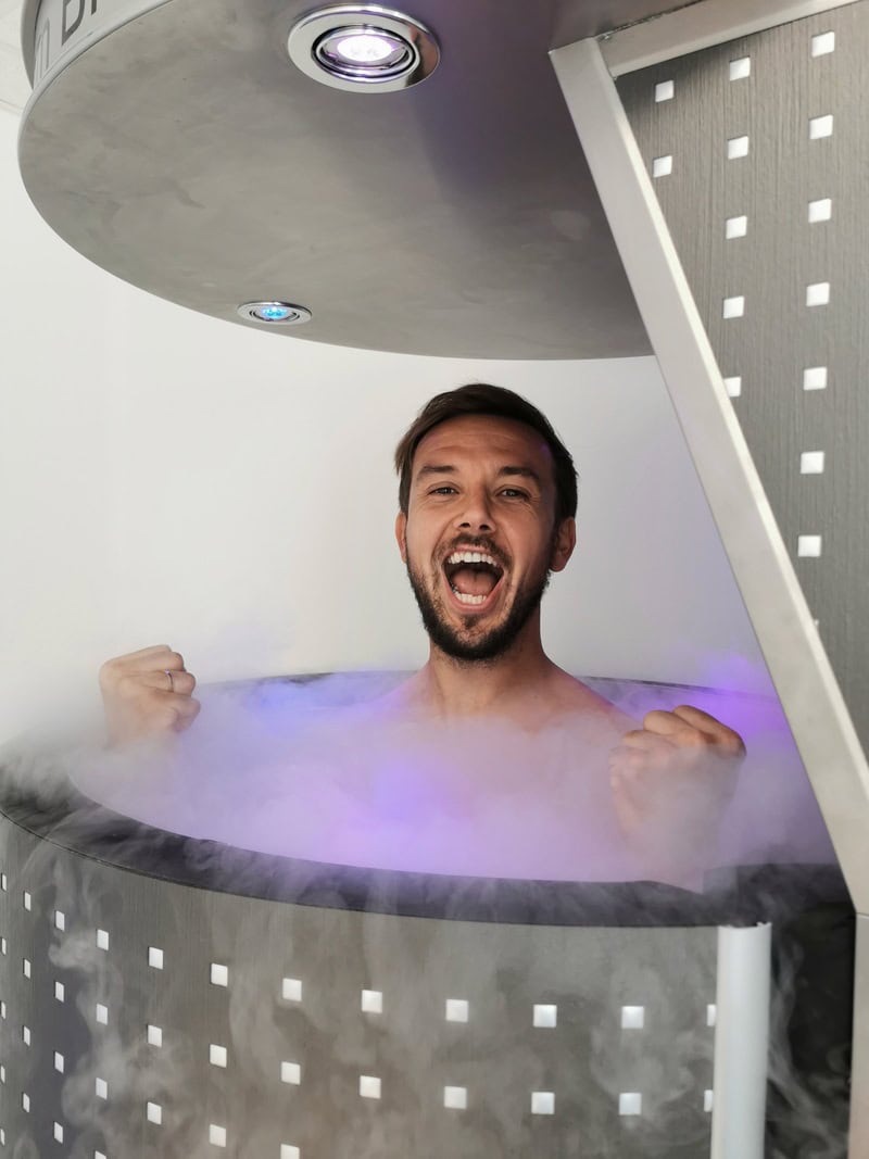 Ein Mann mit freudigem Gesichtsausdruck in einer dampfenden, grauen Kryokammer.