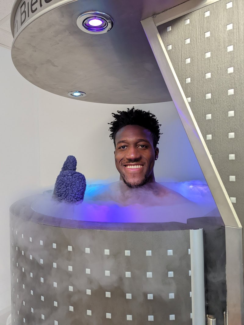 Ein Mann lächelt in einer Kryokammer, trägt einen dunklen Wollhandschuh, umgeben von Nebel.