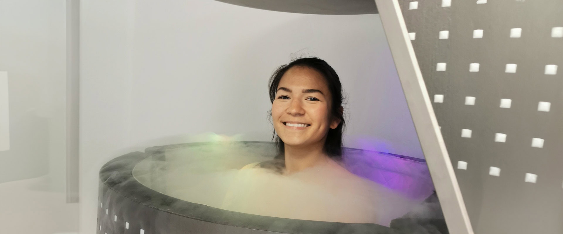 Eine lächelnde Frau in einer Kryokammer mit buntem Dampf und Metallstruktur.