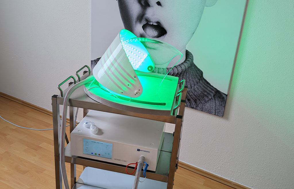 Ein grünes LED-Gesichtsgerät auf einem Metalltisch vor einem stilisierten Porträt.