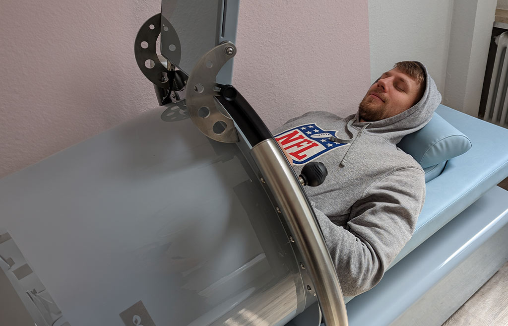 Eine Person entspannt liegend in sportlichem Hoodie auf Blauer Liege mit mechanischem Gerät.