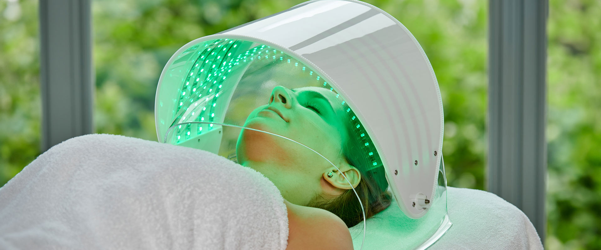 Person in Spa-Behandlung unter grün leuchtender LED-Kuppel, entspanntes Gesicht, weißes Handtuch.
